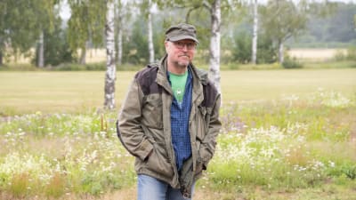 På bilden ser man en man och i bakgrunden finns växter och björjar.