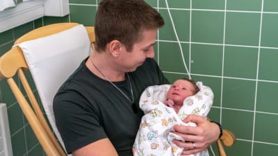 Lucas Lönnqvist sitter i gungstolen och har bebisen i famnen. Bebisen tittar på pappan som ler. 