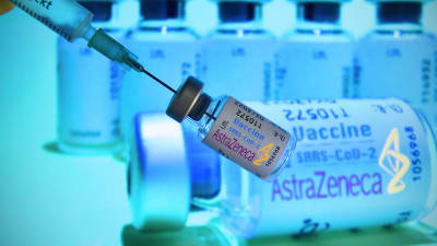 En person håller i en dos med AstraZenecas coronavaccin. I bakgrunden syns fler doser.
