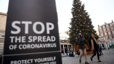 Bild på skylt där det står stoppa spridningen av coronaviruset, och i bakgrunden ett par som går hand i hand vid en stor julgran på julmarknad i London.