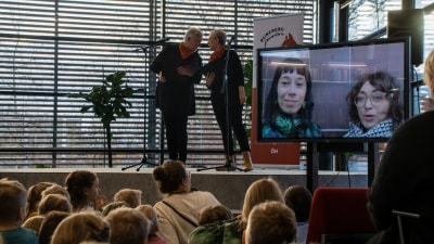 Två kvinnor står på scen och talar med varandra framför en publik med många barn och en del vuxna. Publiken tittar på två kvinnor som syns i en stor tv-skärm.