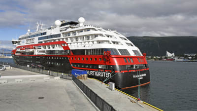 Bild på svart, rött, vitt kryssningsfartyg i hamn. I bakgrunden syns berg.