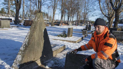 En man i orange arbetskläder sitter på huk och pekar på en gravsten.