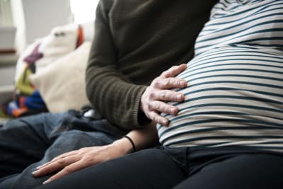 Gravid kvinna sitter på soffa med sin partners hand på magen.