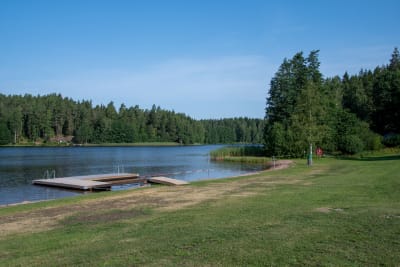 Bild på badstrand med brygga. I änden av stranden finns ett svartgulrandigt band.