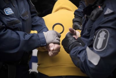 Två poliser sätter handklovar på en person i gul munkjacka.