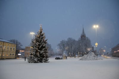 En julgran lyser på ett snötäckt torg. I bakgrunden skymtar en rödtegelkyrka.