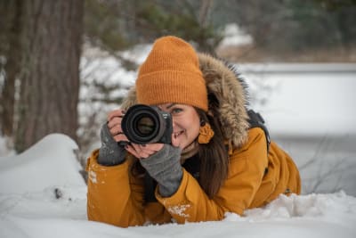 Anette ligger i snön och tar en bild med sin systemkamera. Hon ler. Har en orange jacka och orange mössa. 