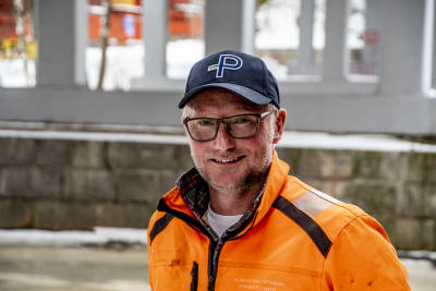 En man i orange jacka, glasögon och keps ler glatt mot kameran