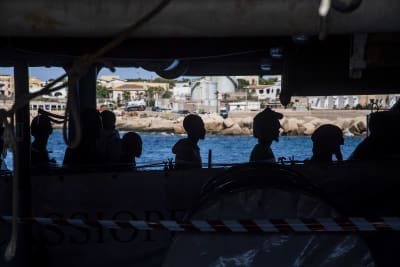 Människosiluetter från sidan, de sitter i en båt med tak. I bakgrunden hav och en stad som ser ut att vara i medelhavsområdet.