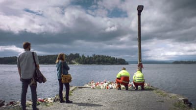 Harald (Marius Lien) och Anine (Alexandra Gjerpen) står vid bryggan mot Utöya, två ambulansmän ställer ner blommor..