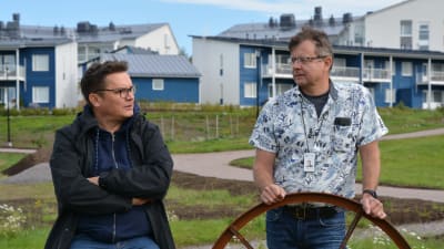 Jarkko Lyytinen och Kari Ojamies står i en park och diskuterar. Blåa hus i bakgrunden.