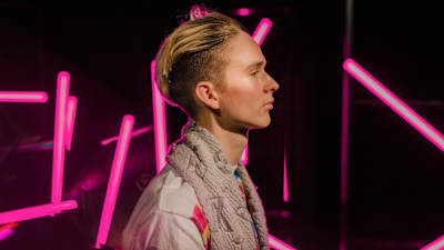 Vaaleahiuksinen mies seisoo sivuttain vaaleanpunaisten neonvalotankojen edessä ilmeettömänä.