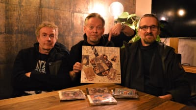 Tre män som håller i en LP-skiva. På bordet framför sig har de fyra cd-skivor.