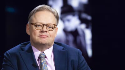 En man klädd i mörkblå kavaj, rosa skjorta och glasögon tittar in i kameran och ler.