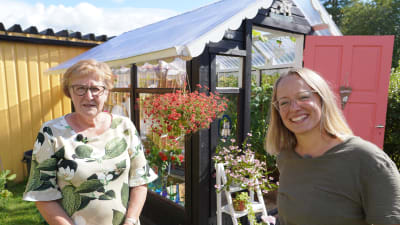 Två kvinnor som står framför ett litet växthus i en blommande trädgård.
