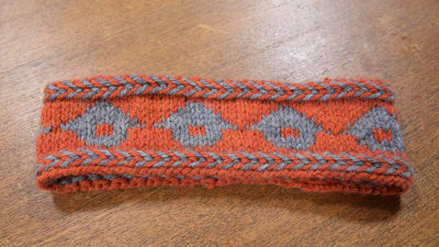Ett mönsterstickat pannband med orange botten och gråa lador.