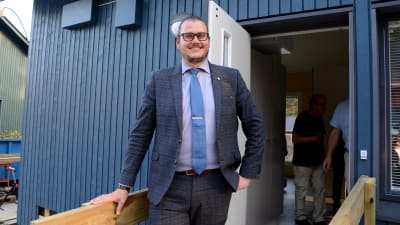 En man står i dörröppningen till ett blått trähus. 