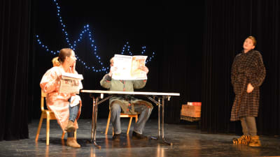 Tre personer på scen. En läser tidning.