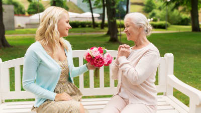 en ung kvinna räcker en blombukett till en äldre dam, båda sitter på en parkbänk. 