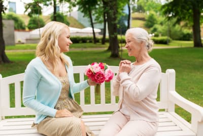 en ung kvinna räcker en blombukett till en äldre dam, båda sitter på en parkbänk. 