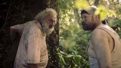 Två ovärdadde män står vid ett stort träd i djungelliknande miljö.