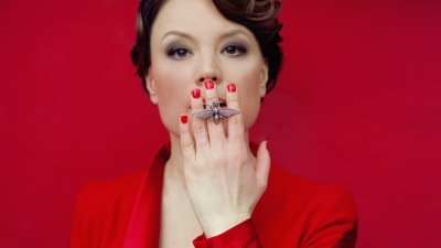 kvinna i klädd röd dräkt håller handen framför munnen. En fjärilsring pryder ett av fingrarna.