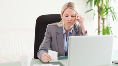 Kvinna sitter bakom en bärbar dator. Hon ser bekymrad ut och ser på en lapp som hon har i handen.