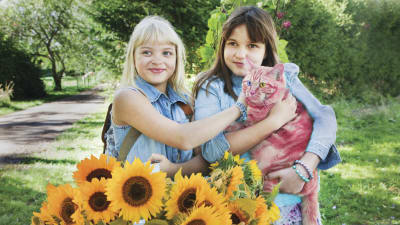 Onneli och Anneli (Lilja Lehto och Aava Merikanto) står invid ett fång med solrosor och håller en röd katt i famnen.