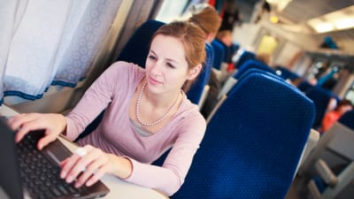 Ung kvinna sitter på ett tåg och skriver på en dator. 