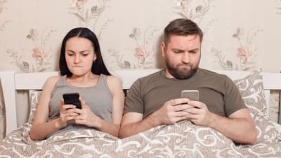 man och kvinna ligger i säng fokuserade på varsin telefon