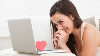 En kvinna sittar framför en laptop med händerna slutna och ser drömskt mot skärmen. Intill skärmen står ett litet hjärta.