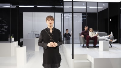 En skådespelare står på en vit scen. I bakgrunden glasväggar och två skådisar som sitter på en soffa. 