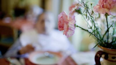 I förgrunden syns en bukett med blommor på ett bord, i bakgrunden syns en äldre kvinna på ett äldre boende som äter soppa.