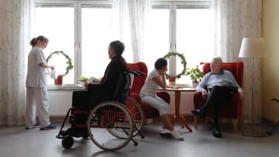 Två personer sitter i var sin fåtölj och samtalar. På bilden finns också en person i rullstol och en person som står vid ett fönster och vattnar en växt.