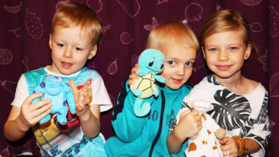 Tre barn, två pojkar till vänster och en flicka till höger, sitter på en bänk framför en lila gardin och håller varsitt kramdjur i handen.