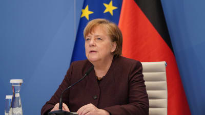 En kvinna i rödaktig jacka sitter med en mikrofon framför sig. Hon har sina händer på bordet. Bakom henne syns två flaggor.
