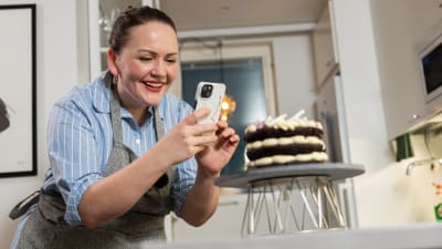 En leende kvinna står och fotograferar en tårta som hon gjort.