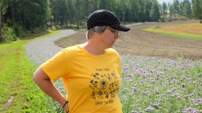 Forskaren Traci Birge tittar ut på ängen i en gul t-tröja med bilder på olika blommor och texten "plant these, save the bees". 