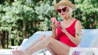 Äldre dam i baddräkt och solglasögon sitter på solstol och tittar leende på telefon 