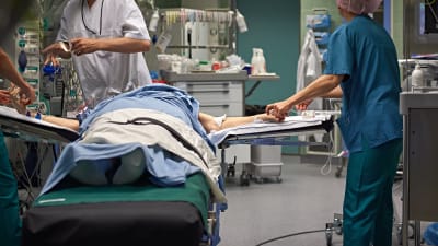 En patient ligger på en säng i en operationssal. Runtom står vårdpersonal.