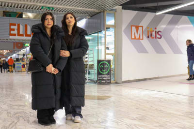 Två unga kvinnor står i armkrok i ett köpcenter. 