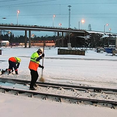 Kaksi miestä harjaa junanraiteita puhtaaksi lumesta.