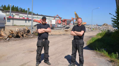 Två män i svarta kläder står framför en tomt där stora maskiner bryter sten