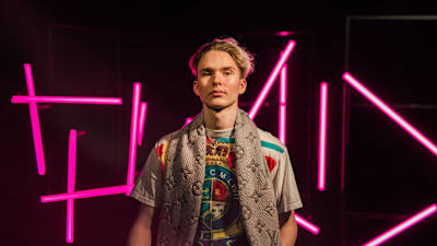 Vaaleahiuksinen nuori mies katsoo kameraan pinkkien neonvalotolppien edessä seisten, kuva rajattu vyötäröstä alaspäin.