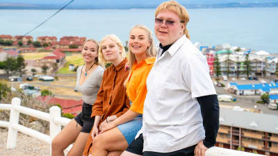 Neljä nuorta au pairia poseeraa iloisina ja värikkäinä merimaiseman edessä.
