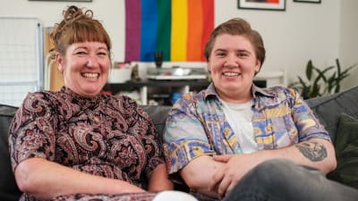 en mamma och hennes som som är trans sitter på en soffa och ler brett till kameran
