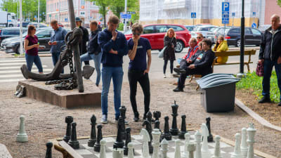Två unga män står och ser fundersamt på ett enormt shackbräde i en park. Runt dem står publik och ser på.