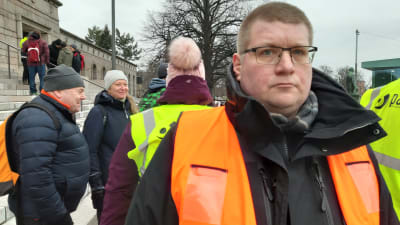 Petteri Syväkari ser bister ut och tittar mot kameran, i orange väst på riksdagshusets trappa, demonstranter i bakgrunden.