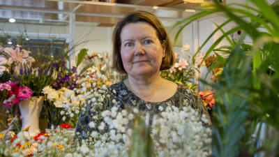 En kvinnlig blomsterhandlare står i sin affär, omgiven av blommor, och tittar in i kameran.